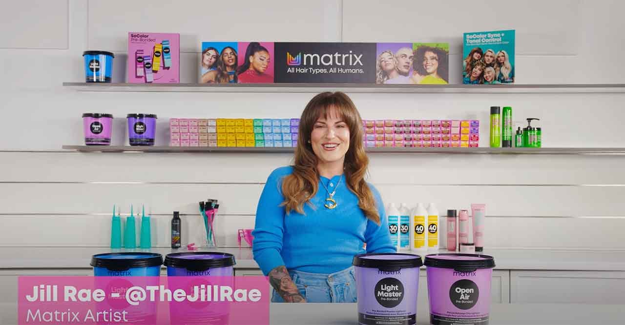 Jill Rae stands in front of a display of Matrix hair products, including Light Master and Open Air lighteners, with colorful hair dye boxes and styling products on shelves behind her. Text reads "Jill Rae - @TheJillRae, Matrix Artist."