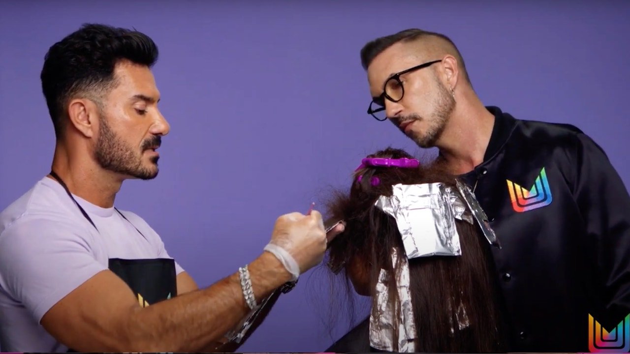 Two hairstylists working on a hair model with long hair. The model's hair is sectioned with foil and clips, and one stylist is applying product while the other observes.