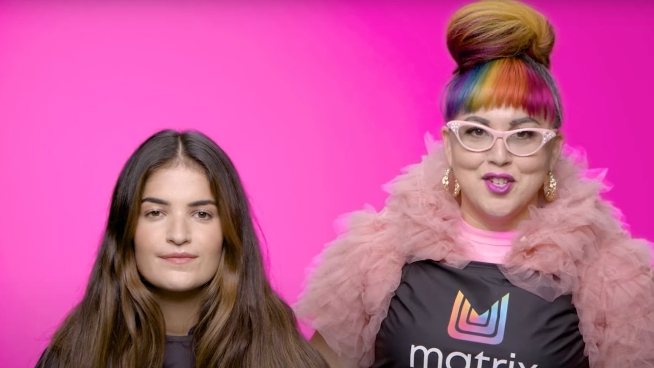 Two individuals in a salon setting with a bright pink background. One is a Matrix stylist who has colorful bangs and is wearing a pink top with ruffled sleeves, while the other is a hair model with long brown hair.