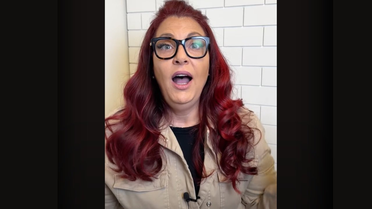 A woman with long, wavy red hair wearing a beige jacket, standing in front of a white tiled wall.